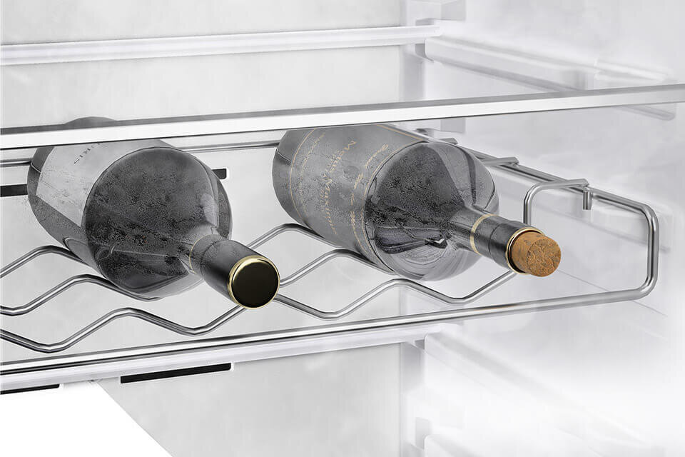 Wnętrze lodówki z widocznym metalowym stojakiem na butelki, na którym leżą dwie butelki wina. Butelki są umieszczone poziomo, a wnętrze lodówki jest jasne i przestronne.