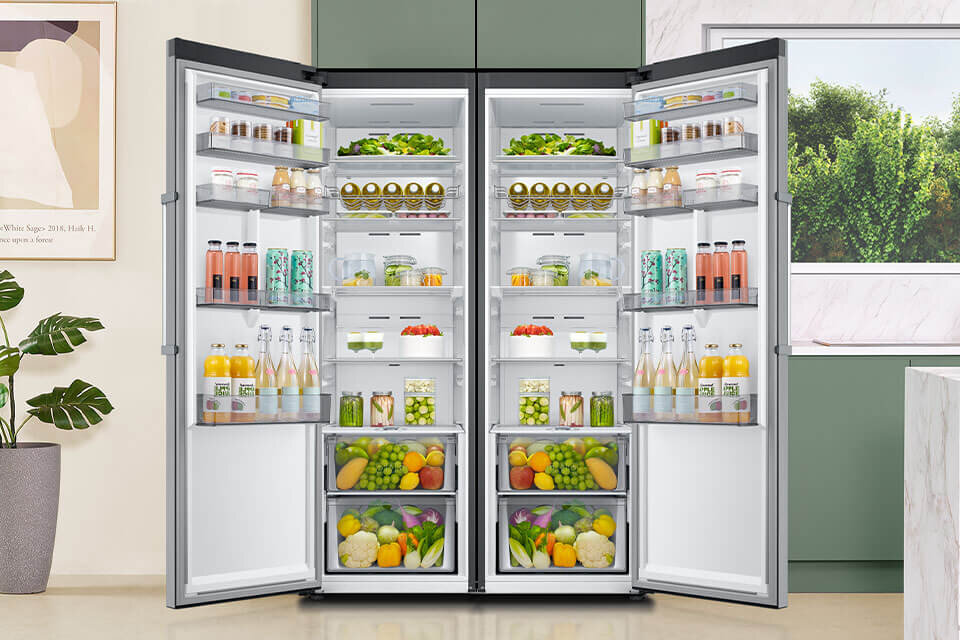 Otwarta lodówka z dwoma drzwiami, wypełniona różnorodnymi produktami spożywczymi, takimi jak owoce, warzywa, napoje i słoiki. W tle widoczne są elementy kuchni, w tym roślina doniczkowa i okno z widokiem na zieleń.