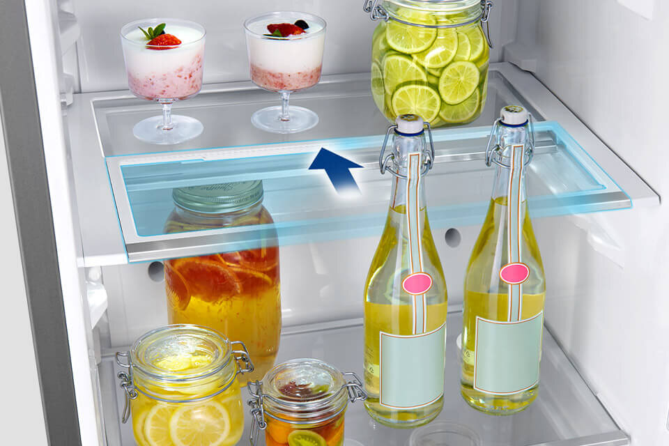 Wnętrze lodówki z półkami, na których znajdują się słoiki z plasterkami cytryny, butelki z napojami oraz deser w pucharkach. Na jednej z półek widoczna jest niebieska strzałka wskazująca na możliwość przesuwania półki.