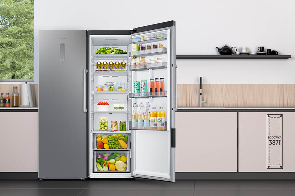 Otwarta lodówka w kolorze inox z widocznymi półkami wypełnionymi różnorodnymi produktami spożywczymi, takimi jak owoce, warzywa i napoje. W tle znajduje się nowoczesna kuchnia z blatem i zlewem, a na szafce widoczny jest napis \'LODÓWKA 3872\'.