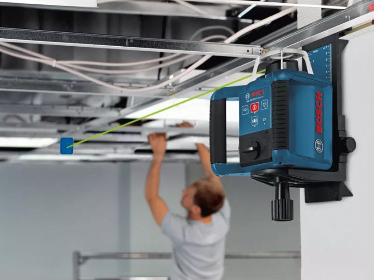 Na obrazie widoczny jest laser rotacyjny z logo BOSCH zamontowany na suficie, emitujący zieloną linię. W tle znajduje się osoba pracująca przy suficie.