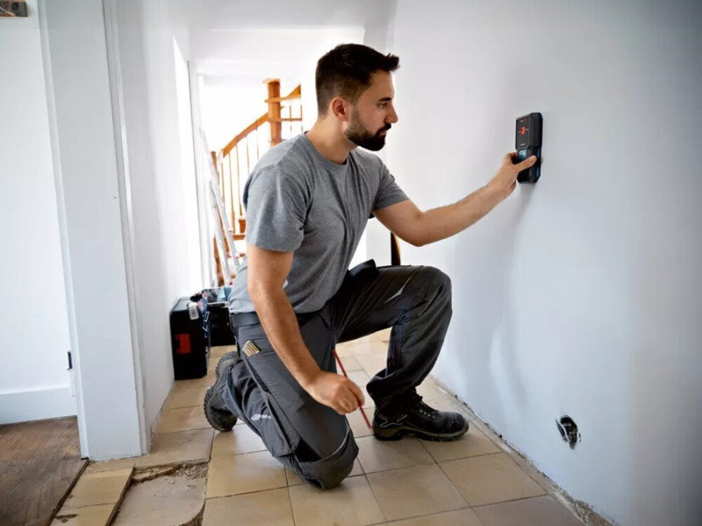 Detektor BOSCH GMS 100-23, dioda, światło, dźwięk, sygnał, mężczyzna przykłada detektor do ściany podczas remontu korytarza