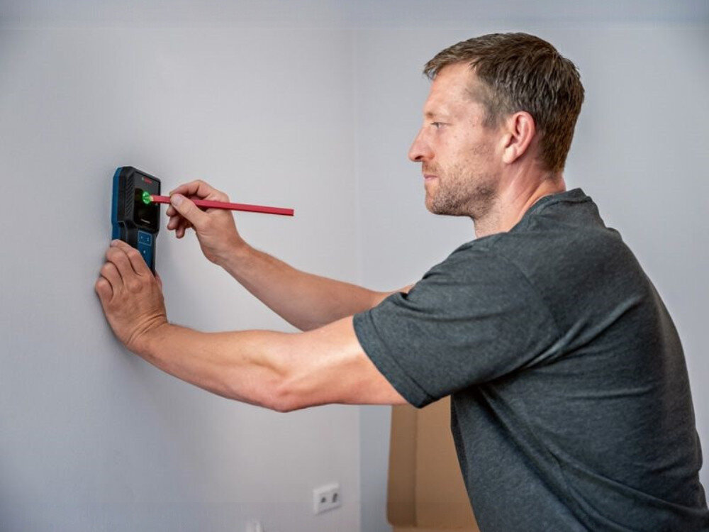 Detektor BOSCH GMS 100-23, ergonomia, waga, wymiary, uchwyt, mężczyzna przykłada detektor do ściany podczas remontu korytarza i znaczy punkt detektorem i ołówkiem