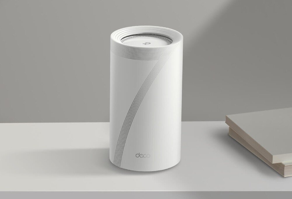 Router TP-LINK Deco BE85 Biały, cylindryczny router z logo 'deco' ustawiony na jasnej powierzchni obok książek, w minimalistycznym wnętrzu, opis
