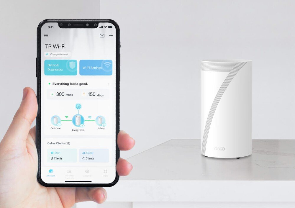 Router TP-LINK Deco BE85 Ręka trzymająca smartfon z aplikacją TP Wi-Fi pokazującą status połączenia sieciowego; obok na blacie stoi urządzenie Deco, aplikacja, narzędzia, kontrola, sterowanie głosem
