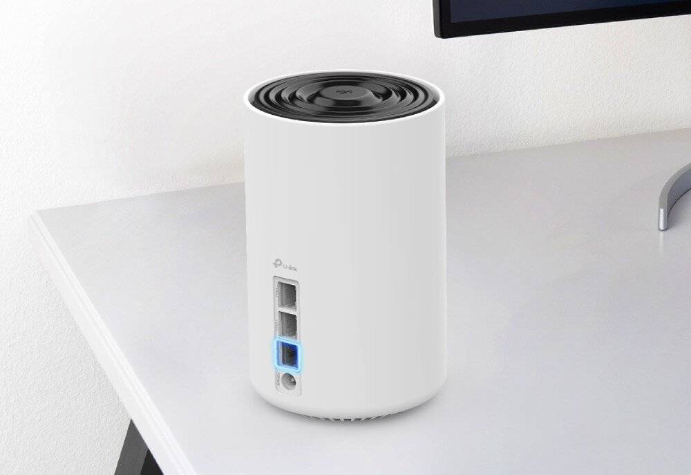 Router TP-LINK Deco XE75 Pro Jedno z urządzeń stojące na biurku obok monitora z widocznymi portami sieciowymi i podświetleniem niebieskiego złącza, złącza Ethernet