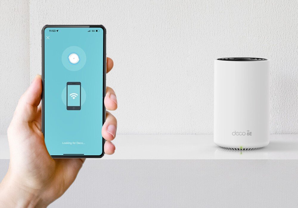 Router TP-LINK Deco XE75 Pro Osoba trzymająca smartfon z aplikacją wyszukującą urządzenie obok stojącego na półce routera z napisem 'deco 6E' i zieloną diodą na dole obudowy, aplikacja, funkcje, narzędzia