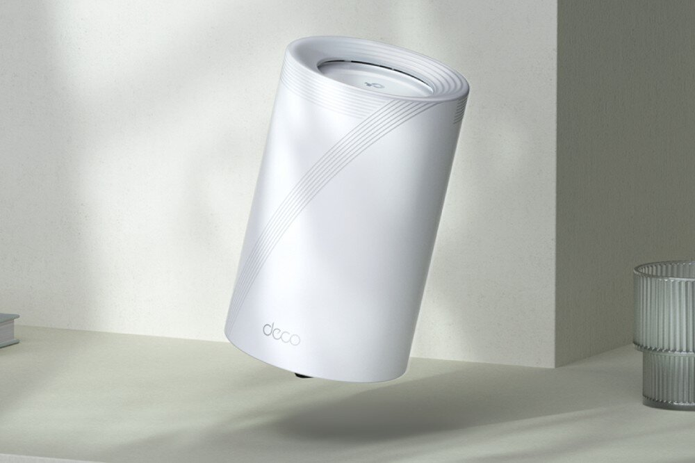 Router TP-LINK Deco BE65 Pojedyncze białe urządzenie w kształcie walca, delikatnie pochylone, znajdujące się na jasnej powierzchni w pobliżu książek i szklanki, opis