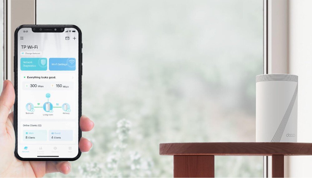 Router TP-LINK Deco BE65 Smartfon trzymany w dłoni, wyświetlający aplikację do zarządzania siecią Wi-Fi. W tle na drewnianym stoliku stoi białe cylindryczne urządzenie, aplikacja, zarządzanie, konfiguracja, sterowanie głosowe