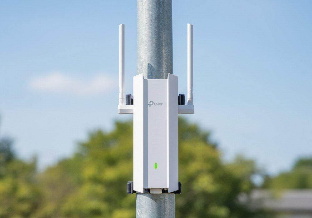 Na obrazie widoczny jest biały punkt dostępu TP-Link z dwoma antenami, zamontowany na metalowym słupie. W tle znajduje się rozmyty krajobraz z drzewami i niebem.