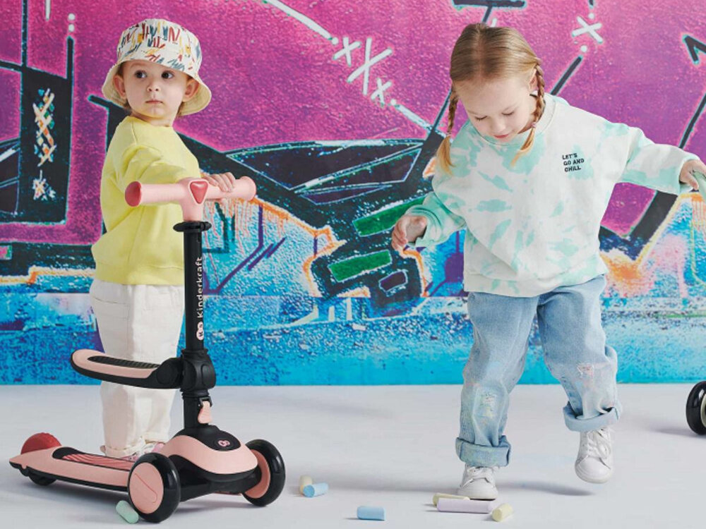 Hulajnoga dla dzieci KINDERKRAFT Halley Różowy dwójka dzieci jedno dziecko opiera się o hulajnogę drugie patrzy w podłogę w tle ściana z kolorowym graffiti aktywność zabawa nauka produkt 2w1