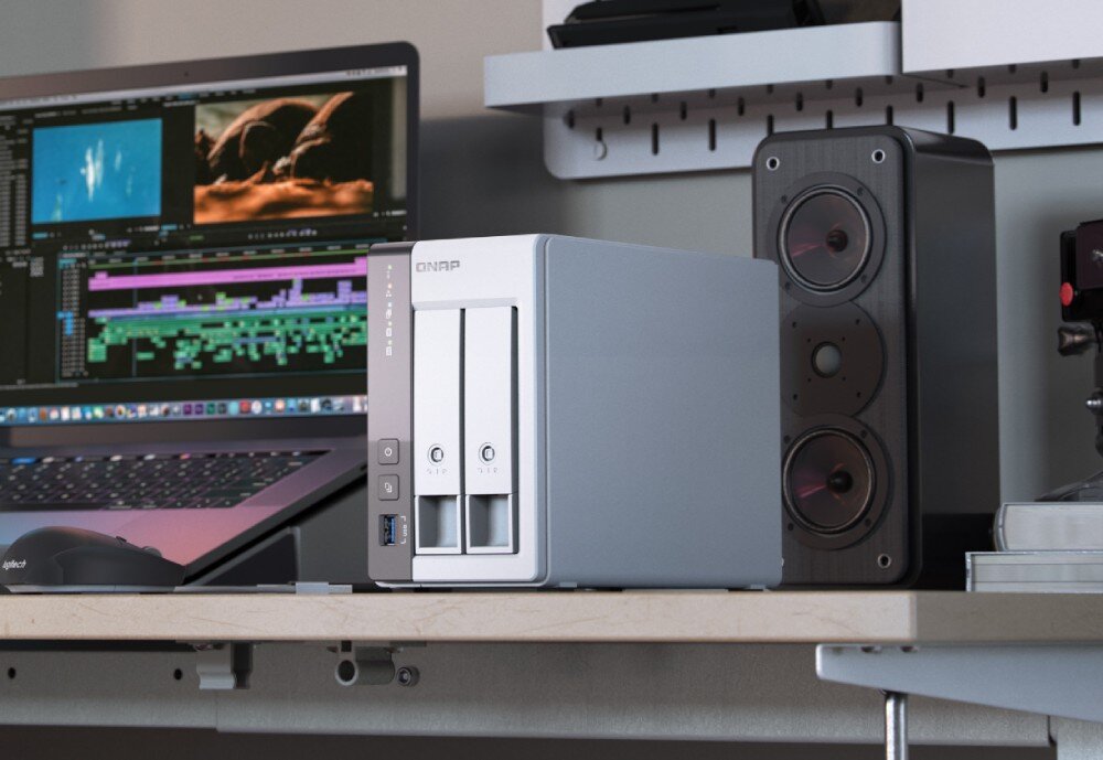 Serwer plików QNAP TS-216G Serwer plików stoi na biurku obok głośnika, a w tle widać otwarty laptop z uruchomionym programem do montażu wideo, opis