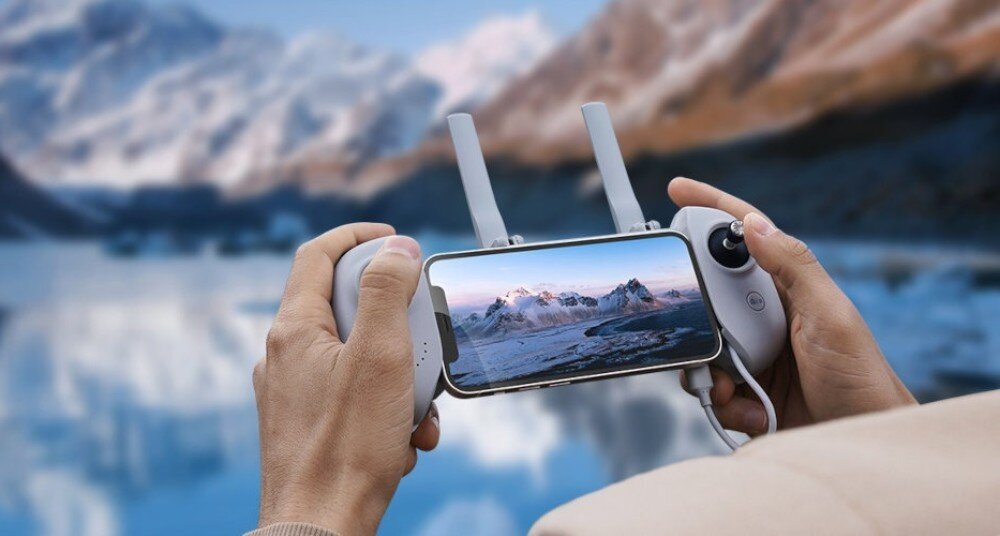 Pilot POTENSIC AT-RC do drona z gimbalem Osoba trzymająca w dłoniach jasnoszary kontroler drona z zamontowanym smartfonem, na ekranie widać obraz z kamery przedstawiający ośnieżone góry i jezioro, w tle rozmyty krajobraz górski