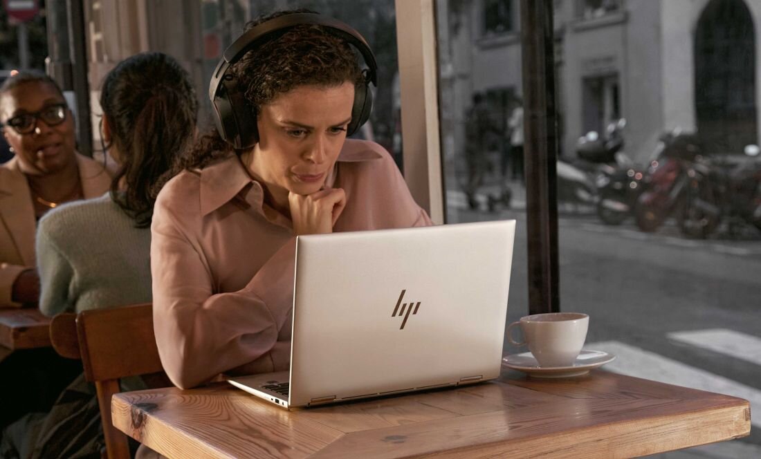 Przy stole siedzi kobieta w słuchawkach, korzystająca z laptopa HP EliteBook 860, zaabsorbowana swoją pracą..
