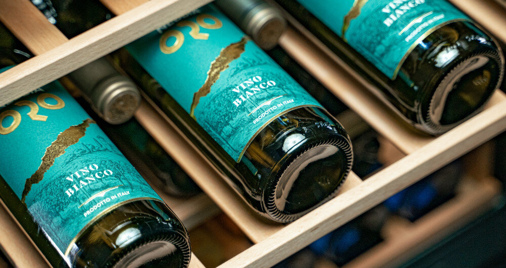 Na drewnianej półce leżą butelki wina z etykietą \'ORO VINO BIANCO\'.