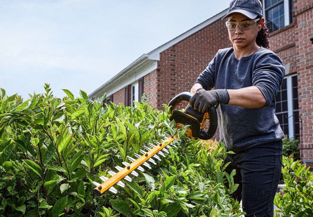 Nożyce do żywopłotu DEWALT DCMHT573N-XJ 650mm 54V akumulatorowe ogród osoba przycina żywopłot w tle dom z cegły niebo pielęgnacja zieleni zadbany ogród sprawne przycinanie żywopłotów krzewów kształtowanie ogrodu