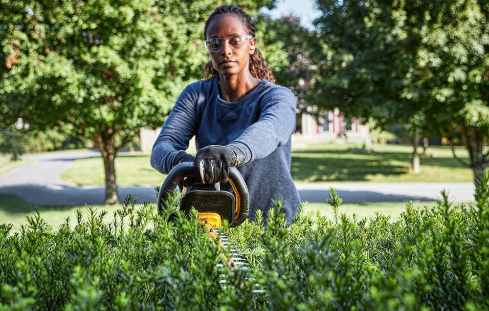 Nożyce do żywopłotu DEWALT DCMHT573N-XJ 650mm 54V akumulatorowe ogród osoba przycina żywopłot w tle drzewa niebo ulica dom do 65 minut na akumulatorze o pojemności 9.0 Ah ciągłość pracy ma realny wpływ na tempo realizacji wygoda użytkowania