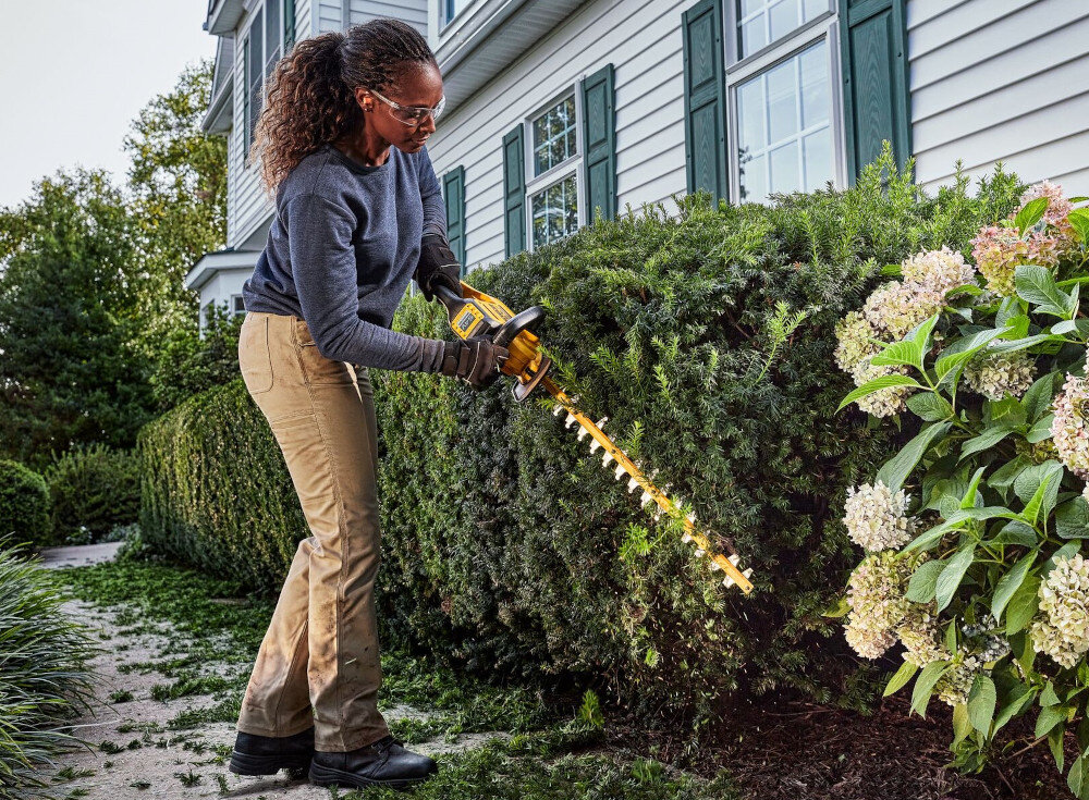 Nożyce do żywopłotu DEWALT DCMHT573N-XJ 650mm 54V akumulatorowe ogród osoba przycina żywopłot przed domem w tle biały dom krzewy kwiaty niebo drzewa dwa przełączniki do uruchamiania nożyc bezpieczeństwo obsługi ogranicza ryzyko przypadkowego włączenia system wymaga świadomego użycia obu rąk
