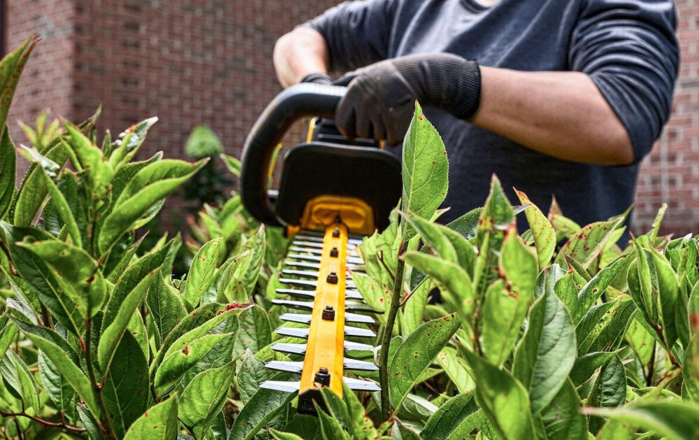 Nożyce do żywopłotu DEWALT DCMHT573N-XJ 650mm 54V akumulatorowe ogród osoba trzyma nożyce w dłoniach w tle dom z cegły uchwyt w kształcie litery D wygodna obsługa w różnych pozycjach dłoni precyzja pracy ogranicza ryzyko nierównego cięcia swoboda zmiany chwytu