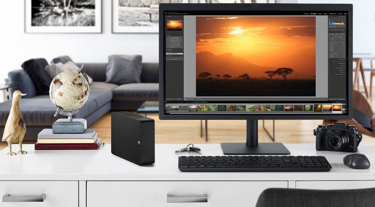 Na biurku znajduje się monitor z wyświetlonym zdjęciem zachodu słońca, klawiatura, mysz, aparat fotograficzny oraz dysk z logo Seagate. Obok monitora stoi globus i kilka książek.