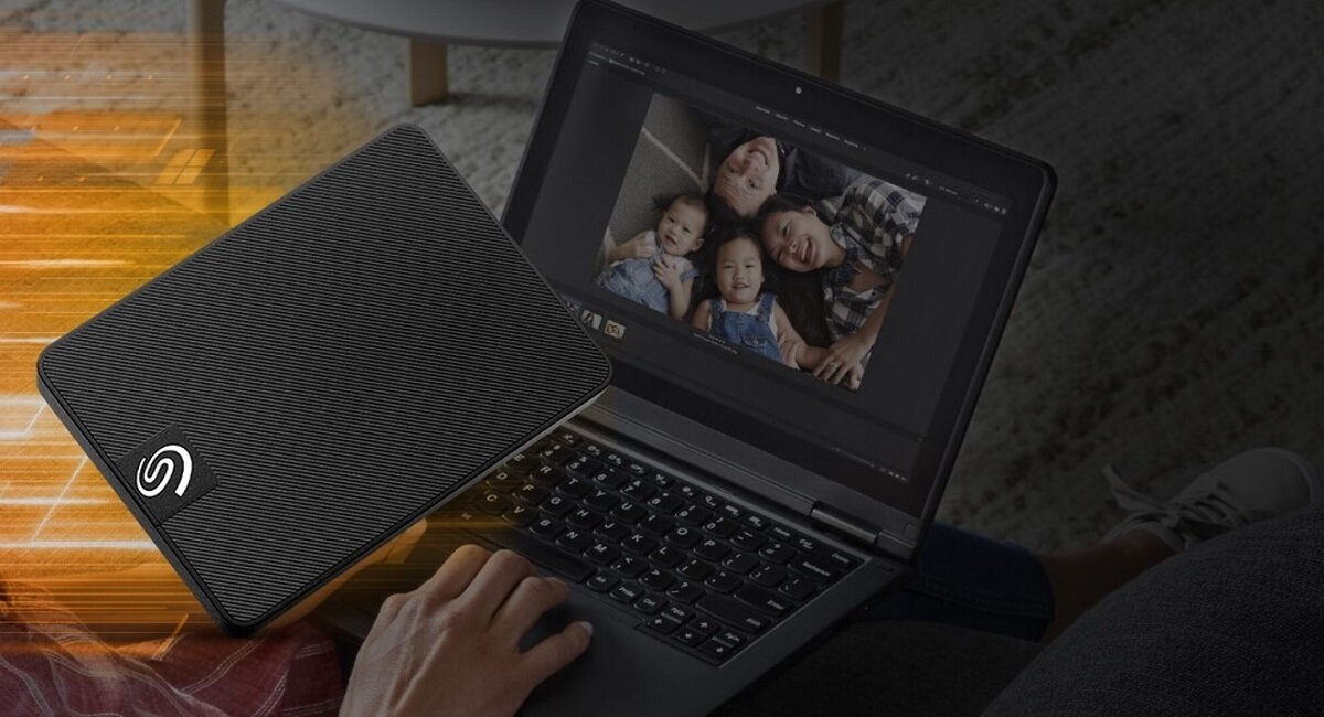 Na zdjęciu widać zewnętrzny dysk twardy z logo Seagate, leżący obok otwartego laptopa, na którym wyświetlane jest zdjęcie rodziny. Dysk ma ciemną, teksturowaną powierzchnię.