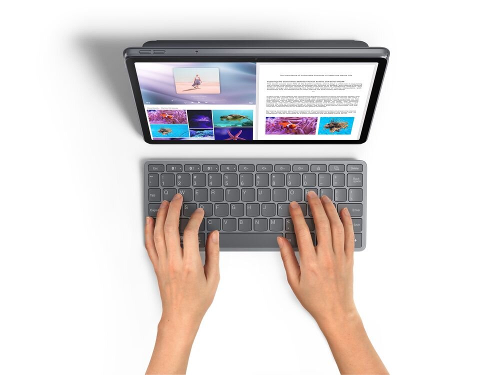 Na obrazie widać tablet z wyświetlonym tekstem i zdjęciami, a przed nim znajduje się klawiatura. Dwie dłonie są ułożone na klawiaturze, gotowe do pisania.