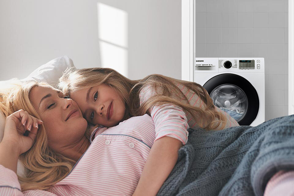 Na obrazie widać pralkę z widocznym logo \'SAMSUNG\' oraz panel sterowania. Obok pralki leżą dwie osoby, które odpoczywają na łóżku.