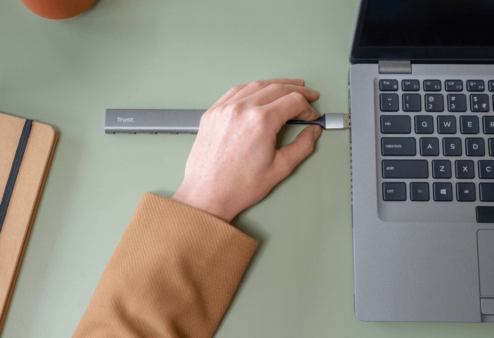 Hub TRUST Halyx 24947 Zbliżenie na rękę w beżowym swetrze wpinającą przewód do portu laptopa z hubem USB widocznym na jasnozielonym blacie