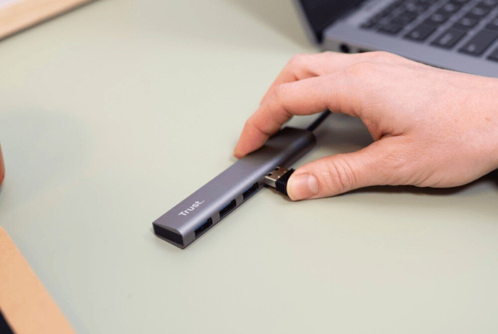 Hub TRUST Halyx 24947 Dłoń podłączająca pendrive do szarego rozdzielacza USB leżącego na białej podkładce biurkowej obok laptopa
