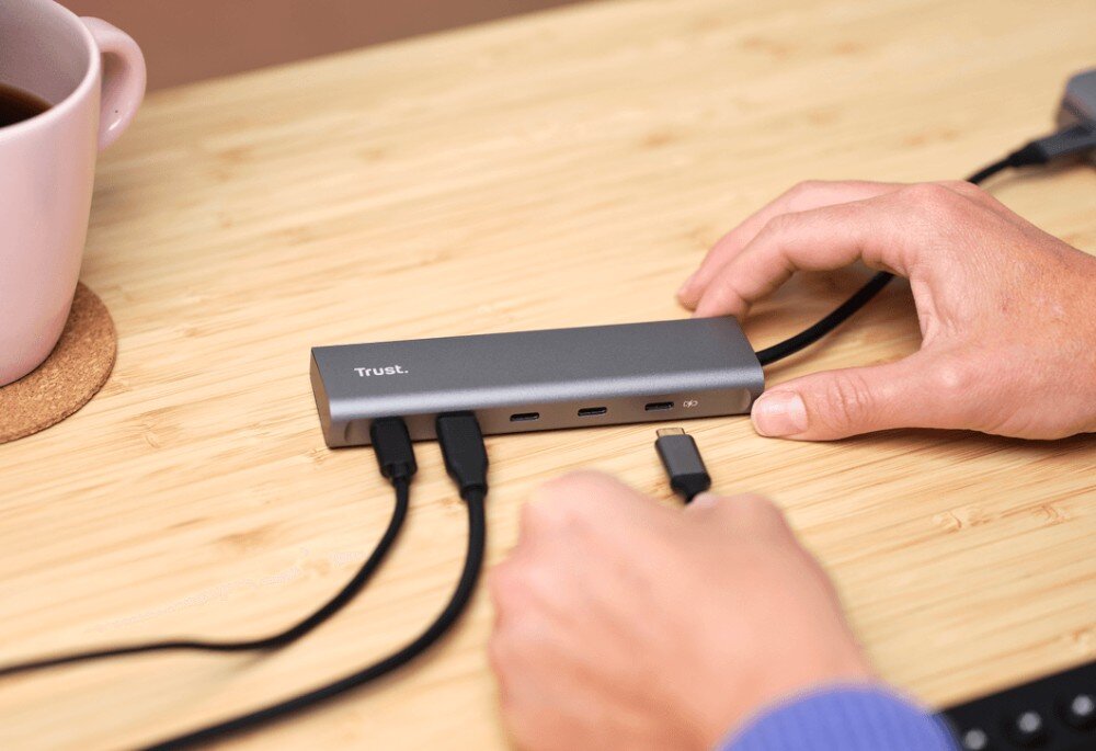Hub TRUST Halyx 25136 Zbliżenie na dłoń podłączającą kabel USB-C do szarego huba leżącego na drewnianym blacie obok różowego kubka