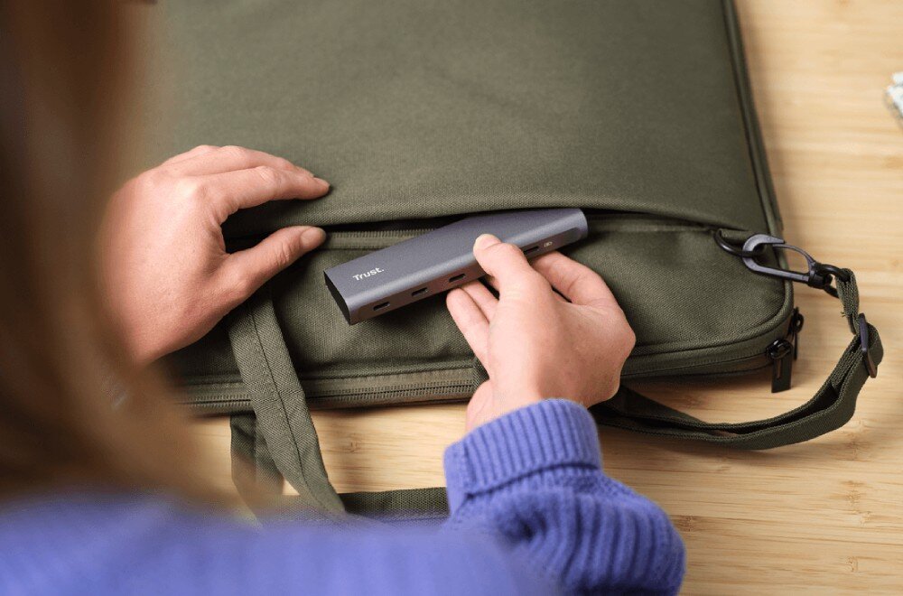 Hub TRUST Halyx 25136 Dłonie w fioletowym swetrze wkładające szary hub USB-C do bocznej kieszeni zielonej torby na laptopa
