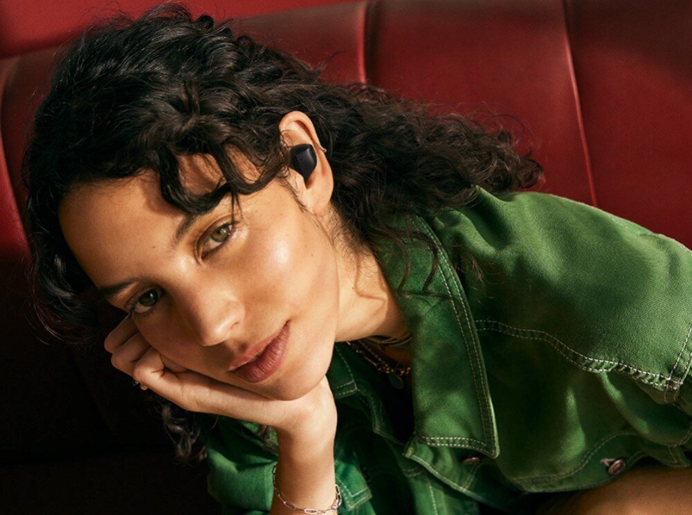 Słuchawki dokanałowe SENNHEISER Accentum True Wireless ANC Młoda kobieta z ciemnymi kręconymi włosami opiera głowę na dłoni, patrząc w stronę aparatu. W jej uszach znajdują się bezprzewodowe słuchawki douszne w czarnym kolorze, opis