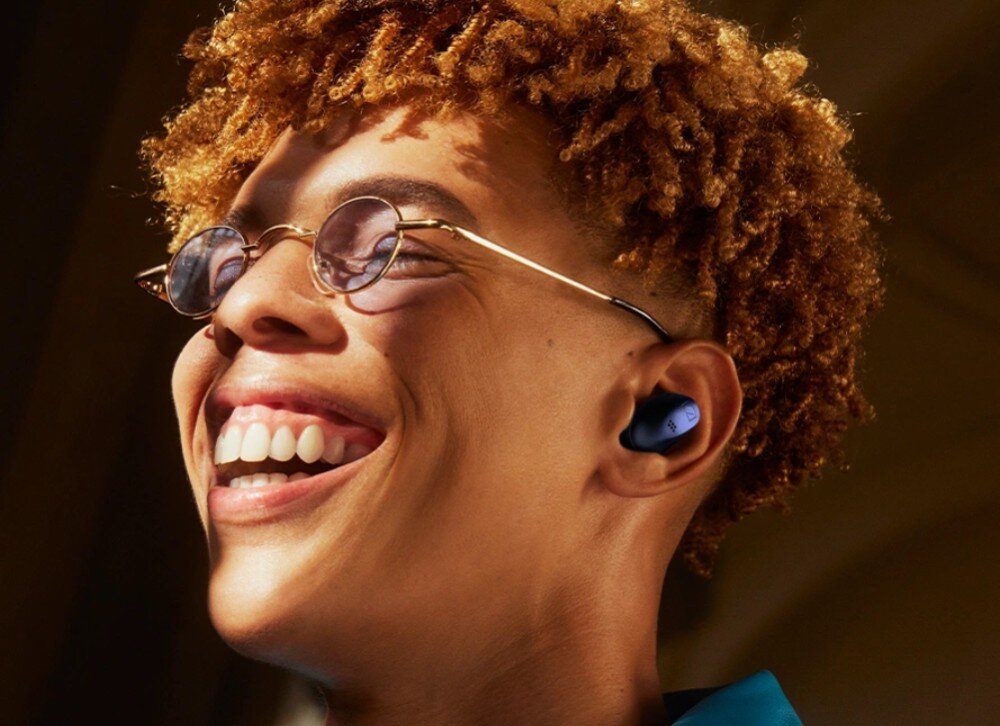 Słuchawki dokanałowe SENNHEISER Accentum True Wireless ANC Mężczyzna z krótkimi, kręconymi rudymi włosami i okularami przeciwsłonecznymi uśmiecha się szeroko. W jego uchu znajduje się czarna bezprzewodowa słuchawka, zalety, cechy, kształt, dopasowanie