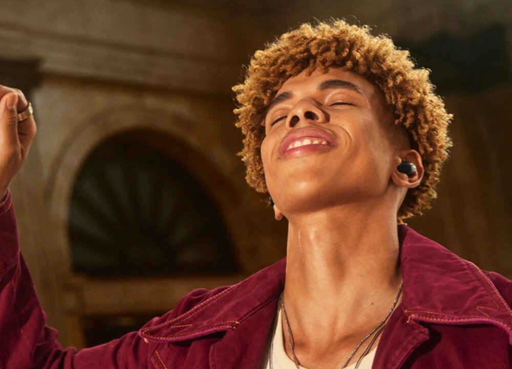 Słuchawki dokanałowe SENNHEISER Accentum True Wireless ANC Mężczyzna z krótkimi kręconymi włosami i szerokim uśmiechem unosi rękę, jakby tańczył lub śpiewał. W jego uszach znajdują się bezprzewodowe słuchawki douszne, łączność Bluetooth, funkcje, sterowanie