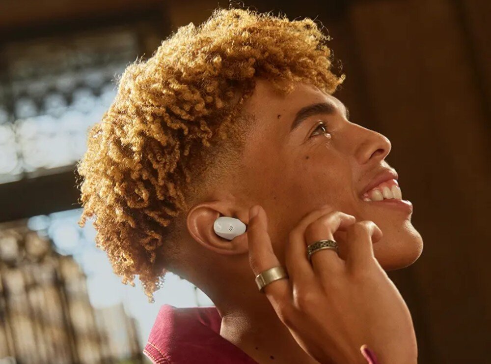 Słuchawki dokanałowe SENNHEISER Accentum True Wireless ANC Mężczyzna z krótkimi, kręconymi rudymi włosami i okularami przeciwsłonecznymi uśmiecha się szeroko. W jego uchu znajduje się czarna bezprzewodowa słuchawka, zalety, cechy, kształt, dopasowanie