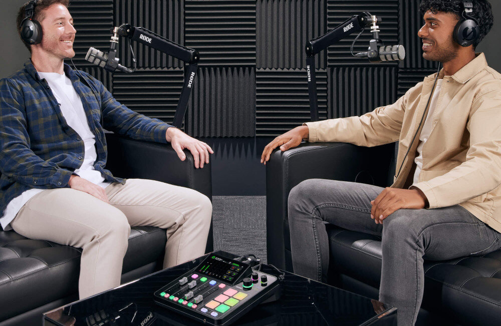 Zestaw RODE Duo Podcast Kit - Zestaw do podcastingu, dwójka ludzi siedzi na fotelach, mając na sobie słuchawki i mówiąc do mikrofonów na statywach, na stoliku leży interfejs