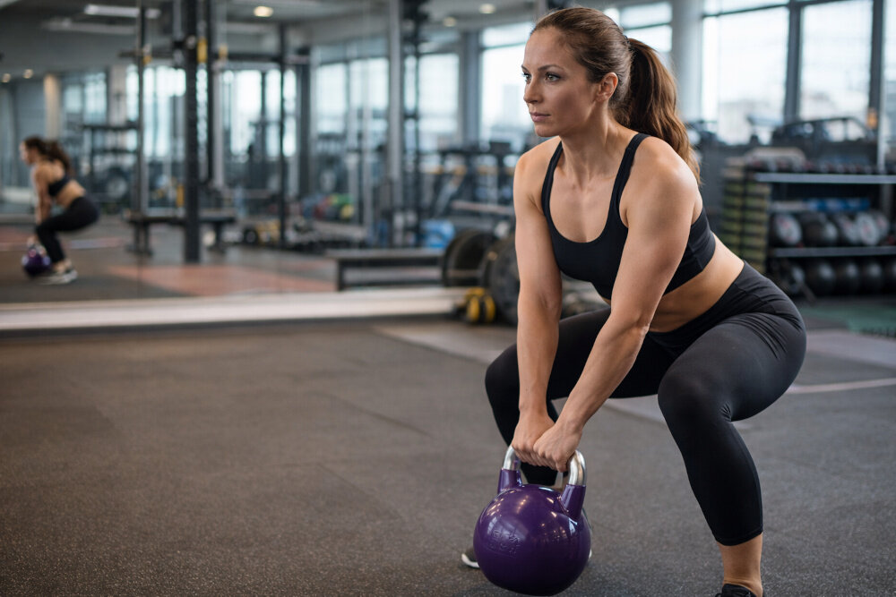 Kettlebell GORILLA SPORTS 2906 (20 kg) sala gimnastyczna siłownia osoba w stroju sportowym robi przysiady trzyma w dłoniach kettlebell trening siłowy kondycja zdrowie wszechstronne zastosowanie