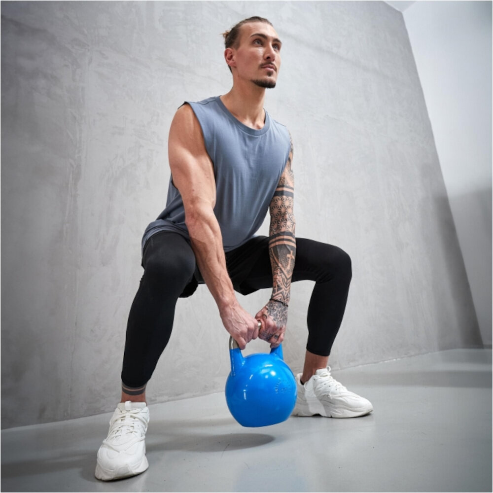 Kettlebell GORILLA SPORTS 2906 (20 kg) szare tło osoba w stroju sportowym wykonuje przysiad z kettlebellem w dłoniach aktywuje mięśnie głębokie odpowiedzialne za stabilizację kręgosłupa wzmacnia wytrzymałość mięśni poprawia postawę ciała równoczesny rozwój kilku grup mięśniowych lepsza kondycja większa kontrola nad ciałem atrakcyjniejszy wygląd sylwetki
