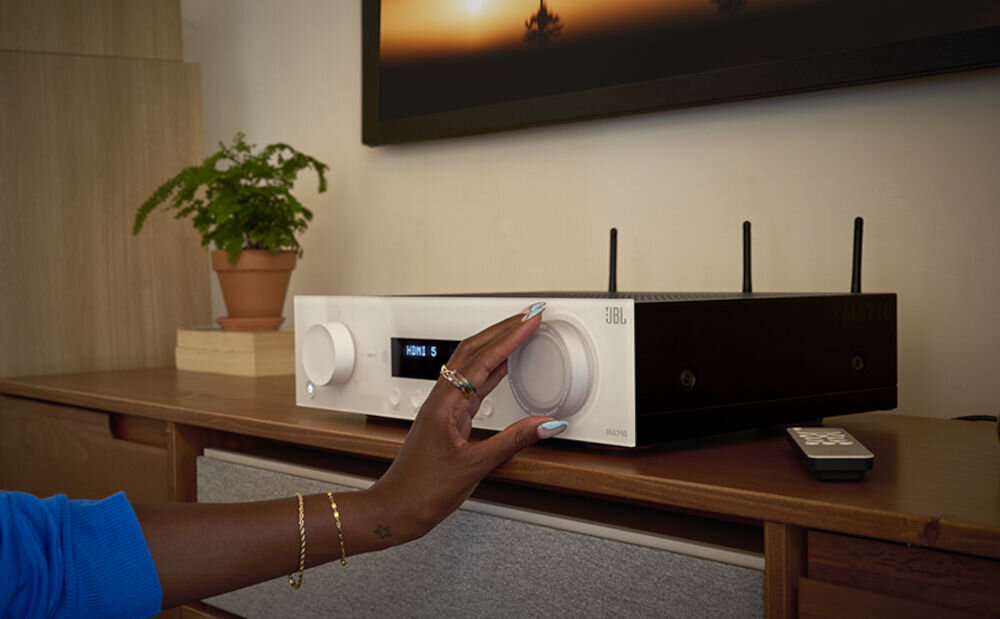 Amplituner JBL MA710 integracja HDMI Smart TV konsole dekodery, osoba dotykająca pokrętła amplitunera stojącego na komodzie