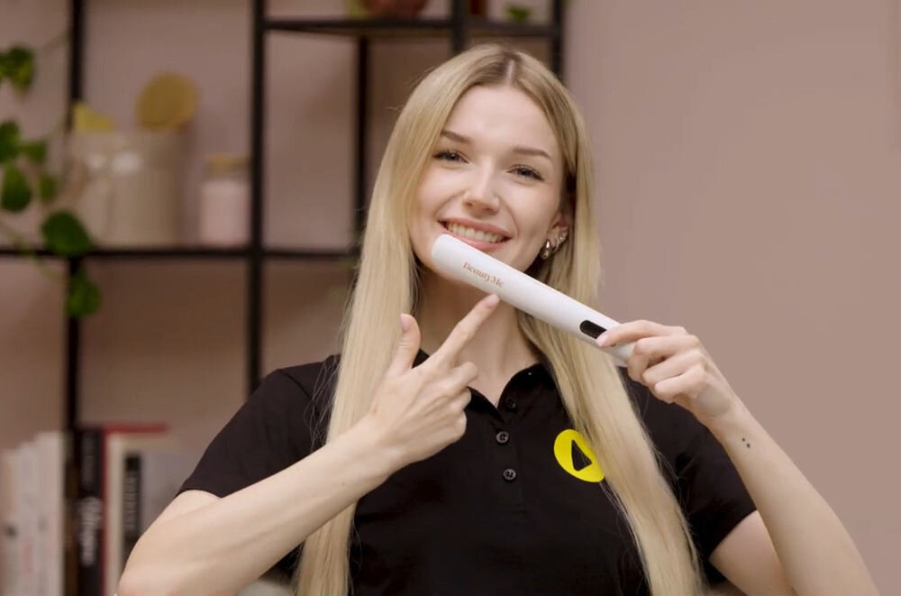 Prostownica bezprzewodowa BEAUTYME HS707W Uśmiechnięta blondynka trzyma w dłoniach bezprzewodową prostownicę BeautyMe HS707W, wskazując palcem na jej obudowę w profesjonalnie urządzonym wnętrzu.
