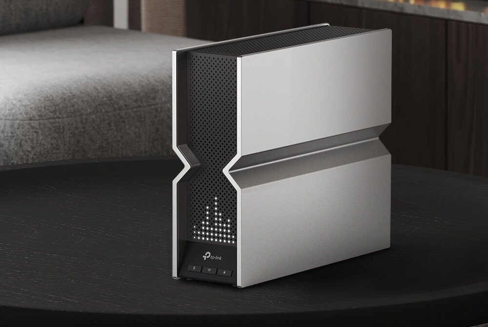 Router TP-LINK Archer BE800 Obraz przedstawia router ustawiony na czarnym stole. Obudowa urządzenia ma srebrny odcień, a na ekranie LED wyświetlany jest pikselowy wzór, obudowa wygląd