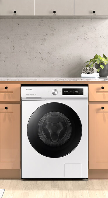 Pralka w zabudowie kuchennej z widocznym panelem sterowania i dużym okrągłym bębnem. Na blacie obok pralki stoi roślina w doniczce i złożone ręczniki.