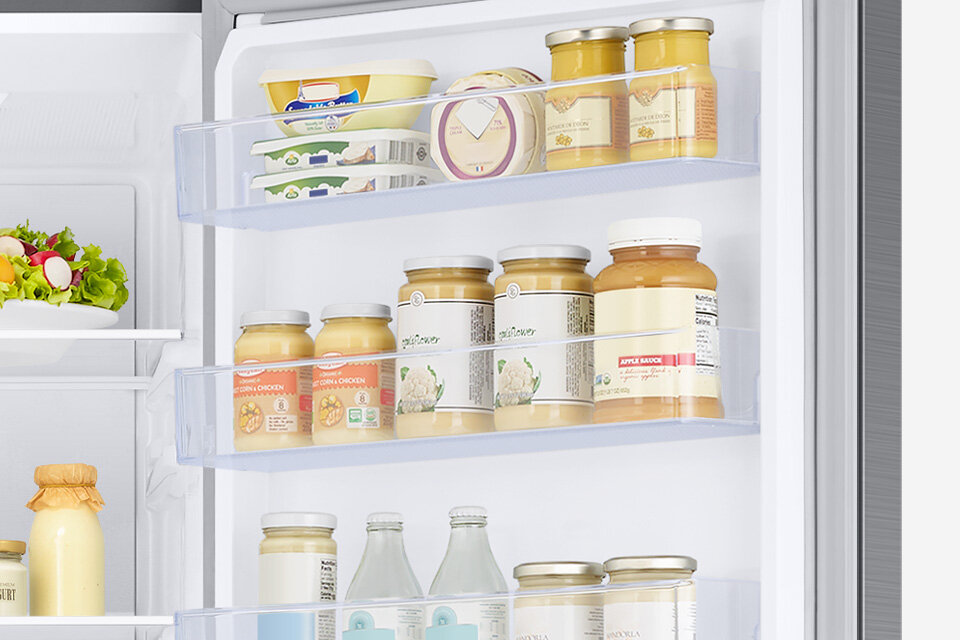 Wnętrze lodówki z widocznymi półkami na drzwiach, na których znajdują się słoiki z przetworami, butelki oraz opakowania z nabiałem. W tle widać część wnętrza lodówki z sałatką na półce.