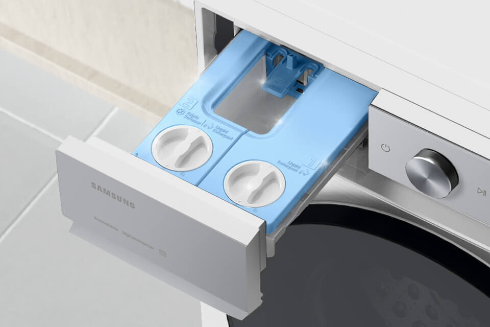 szuflada pralki Samsung, w której nie pozostają osady po detergentach Zdjęcie przedstawia otwarty pojemnik na detergenty w pralce z widocznymi dwoma okrągłymi pokrętłami. Na froncie pojemnika znajduje się logo \'SAMSUNG\'.