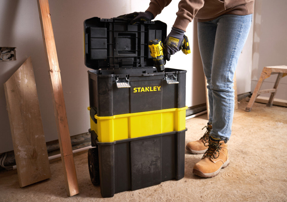 Wózek narzędziowy z organizerem STANLEY STST1-81320 pomieszczenie drewniana podłoga skrzynki ustawione na podłodze osoba wyciąga z wnętrza wiertarkę drewniana ściana drewniana podłoga ruchomy warsztat modułowość mobilność transparent i przechowywanie narzędzi