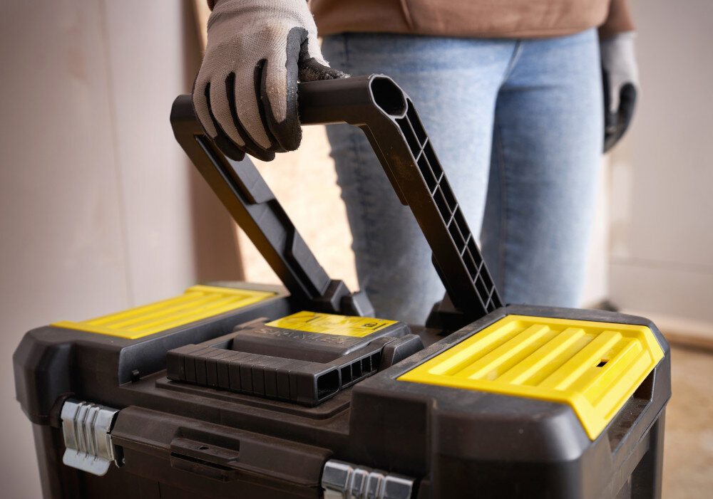 Wózek narzędziowy z organizerem STANLEY STST1-81320 pomieszczenie osoba trzyma uchwyt w dłoni podnosi go kółka teleskopowy uchwyt łatwe przemieszczanie stabilna konstrukcja ogranicza wysiłek ułatwia organizację pracy