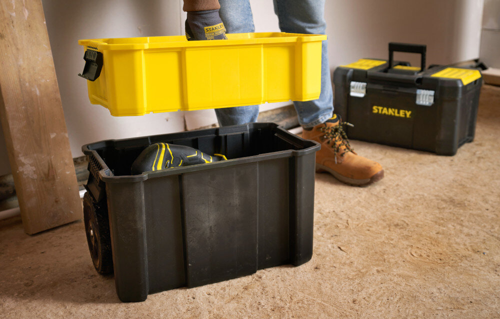 Wózek narzędziowy z organizerem STANLEY STST1-81320 pomieszczenie osoba trzyma żółtą skrzynkę w dłoni na podłodze stoją dwie skrzynki drewniana podłoga wymiary 630 × 475 × 475 mm waga około 4,4 kg stabilność metalowe zatrzaski solidne uchwyty Bi-Materiał pewne przenoszenie skrzynek bezpieczeństwo wygoda codziennej pracy