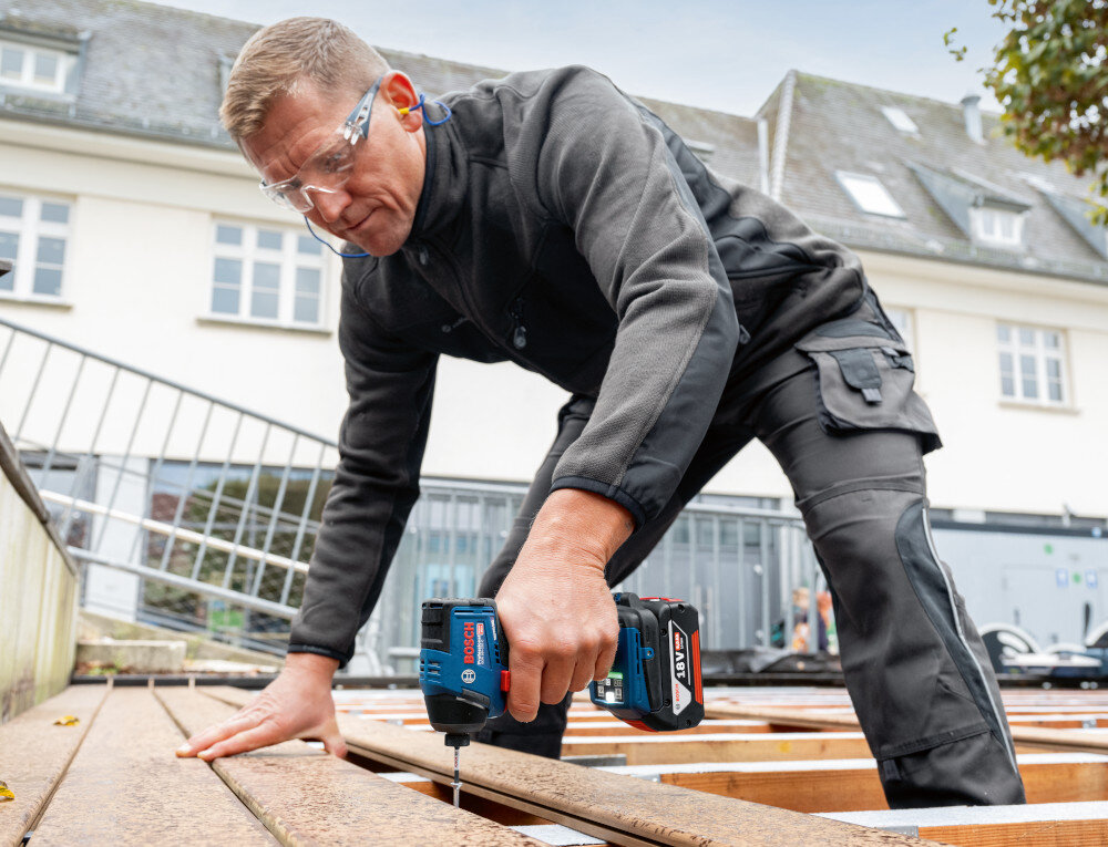 Klucz udarowy BOSCH Professional GDR 18V-220 C 06019L6000 podwórko osoba przykręca śrubę do drewnianego podestu w tle biały budynek do wkręcania i wykręcania śrub wkrętów w drewnie, metalu i konstrukcjach instalacyjnych oszczędza czas