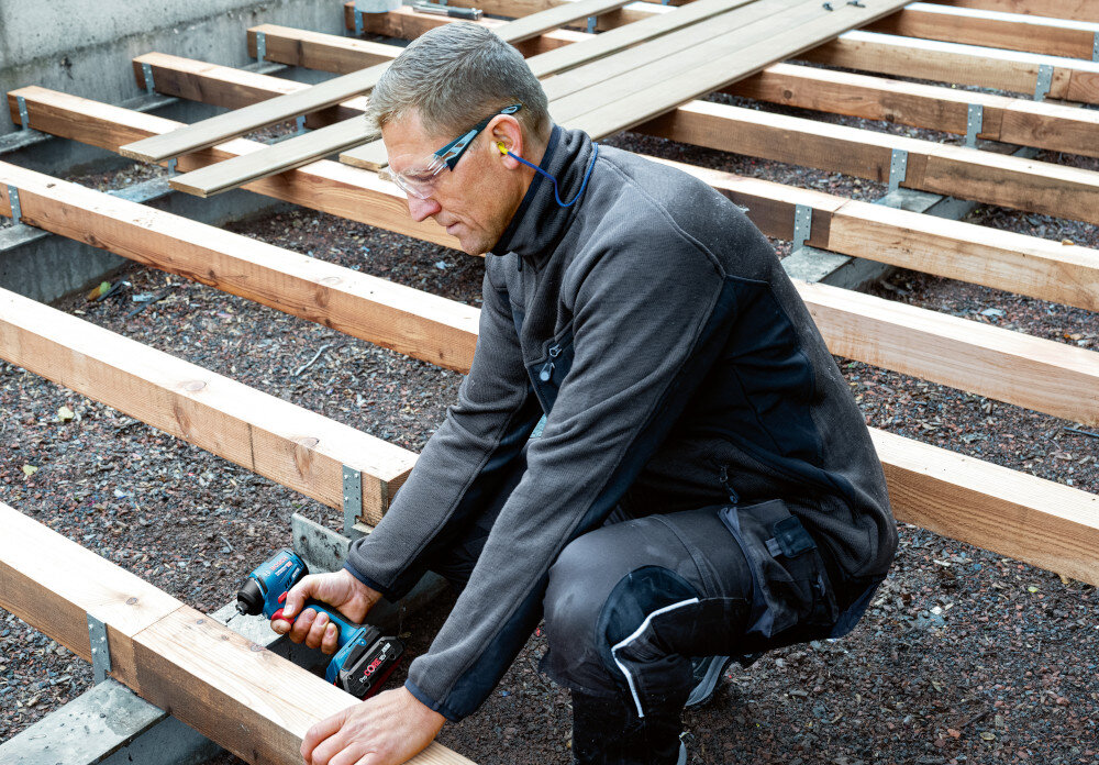 Klucz udarowy BOSCH Professional GDR 18V-220 C 06019L6000 podwórko osoba dokręca śrubę do drewnianej belki podest deski ziemia metalowy zaczep można wygodnie zawiesić na pasku, narzędziowym pasie roboczym lub drabinie ogranicza się konieczność odkładania urządzenia na powierzchnię zwiększa komfort i tempo pracy