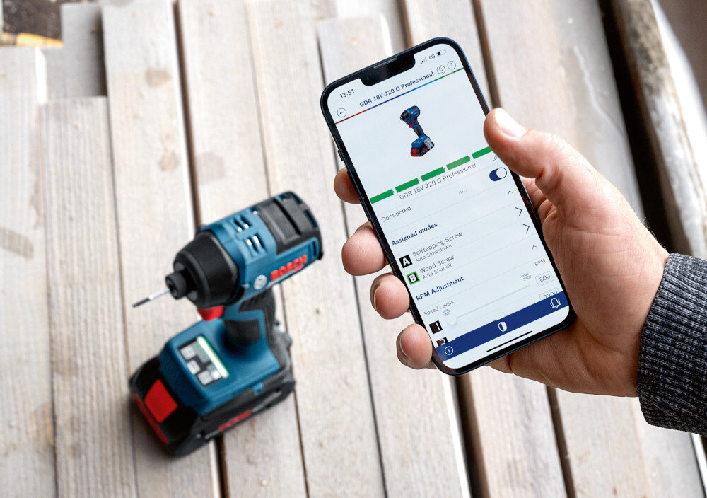 Klucz udarowy BOSCH Professional GDR 18V-220 C 06019L6000 drewniany podest na podeście stoi klucz na pierwszym planie dłoń osoby z telefonem w dłoni włączona aplikacja regulowane tryby wkręcania dostosowanie pracy narzędzia do rodzaju materiału i średnicy wkrętu tryby spowalniają (A) wyłączają (B) napęd zmniejsza ryzyko złamania łbów wkrętów i uszkodzenia obrabianych elementów aplikacja BOSCH Toolbox moduł Bluetooth Connectivity (brak w zestawie)